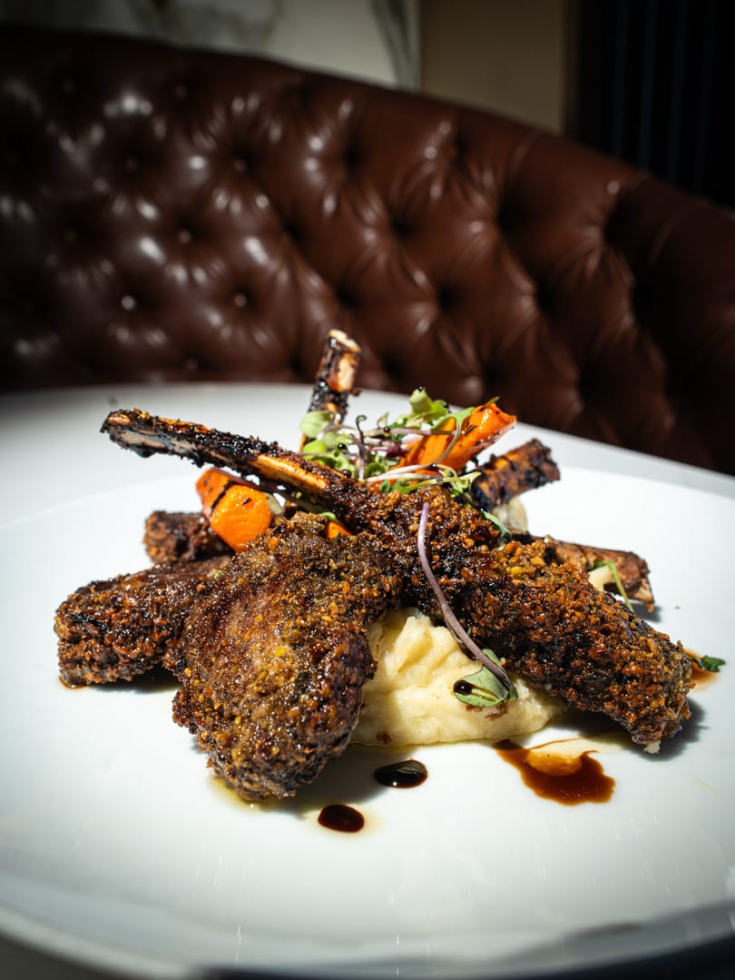 Crispy lamb chops with mashed potatoes, roasted carrots, and microgreens on a white plate.
