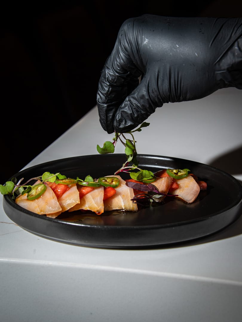 A gloved hand garnishes a black plate of salmon sashimi with microgreens, jalapeños, and red sauce.