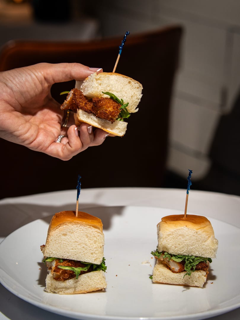 A hand holds a mini fried chicken slider above two more on a white plate.
