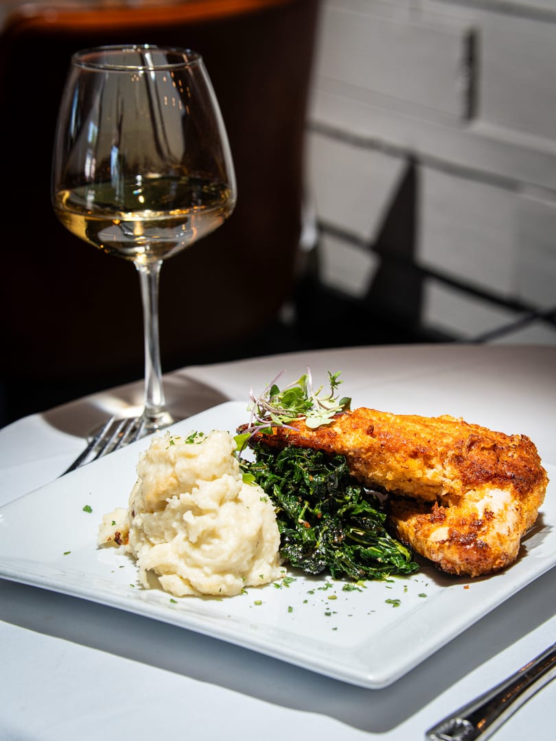 Crispy fried chicken, mashed potatoes, and sautéed greens on a white plate with a glass of white wine.