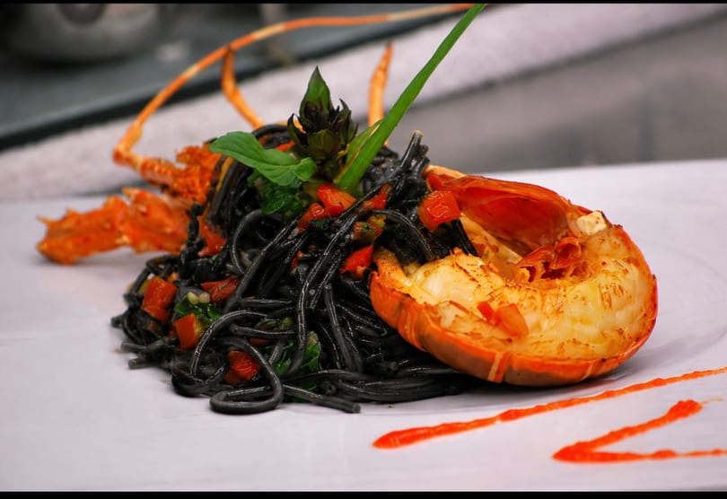 Black squid ink pasta with diced tomatoes, basil, and a grilled lobster tail on a white plate.
