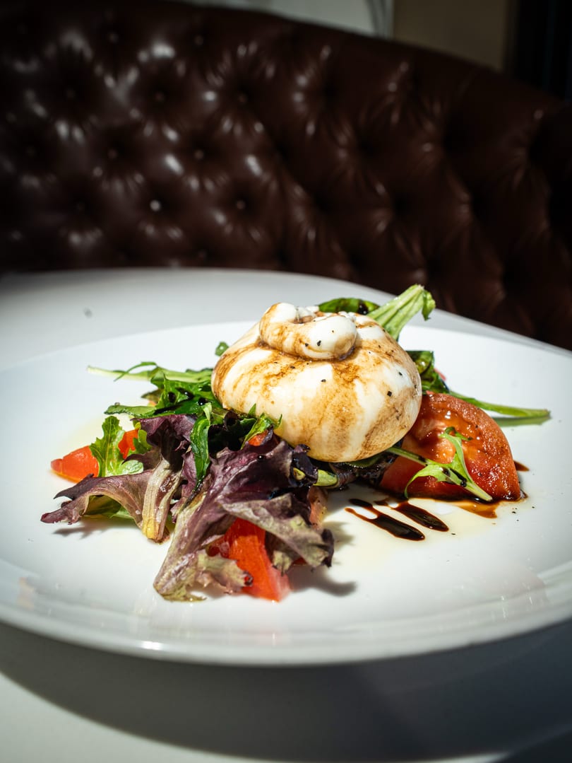Burrata cheese with balsamic glaze, mixed greens, and sliced tomatoes on a white plate.