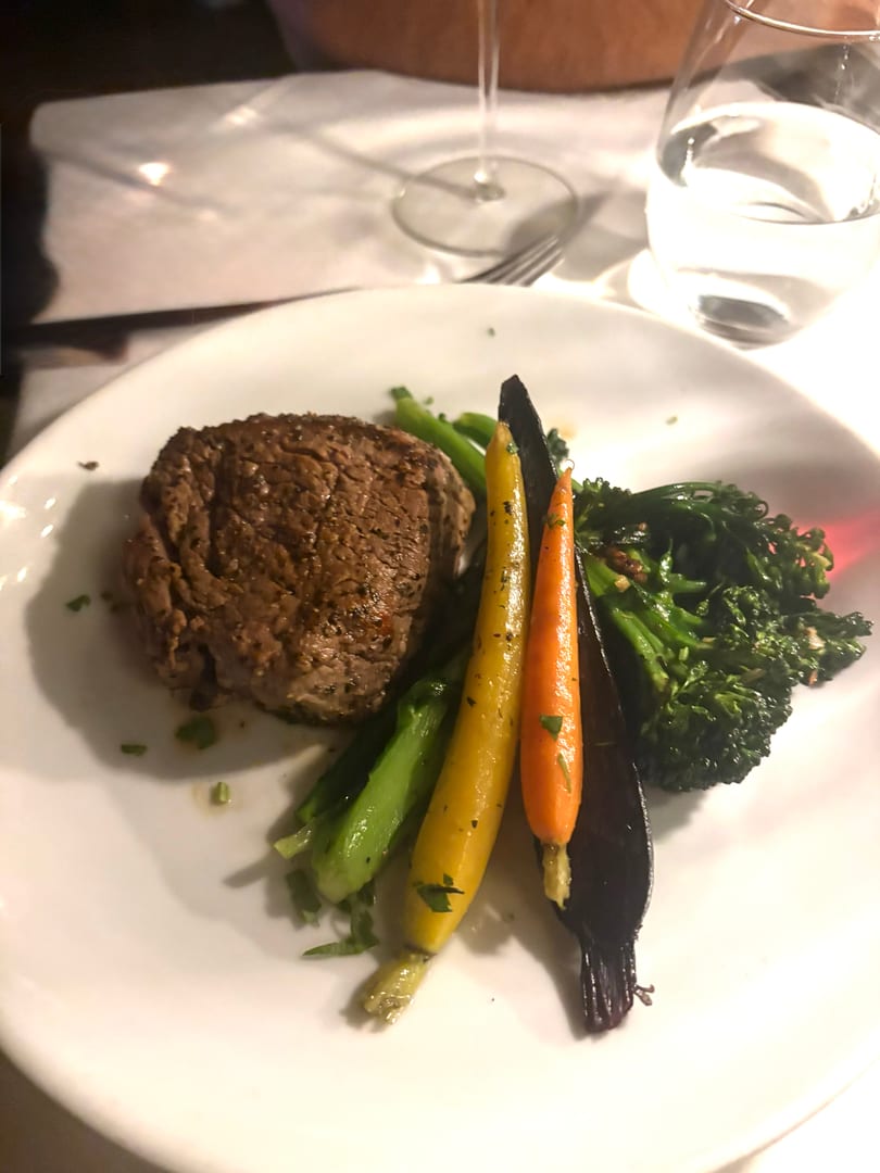 A steak entree with broccolini and colorful carrots on a white plate at a restaurant.