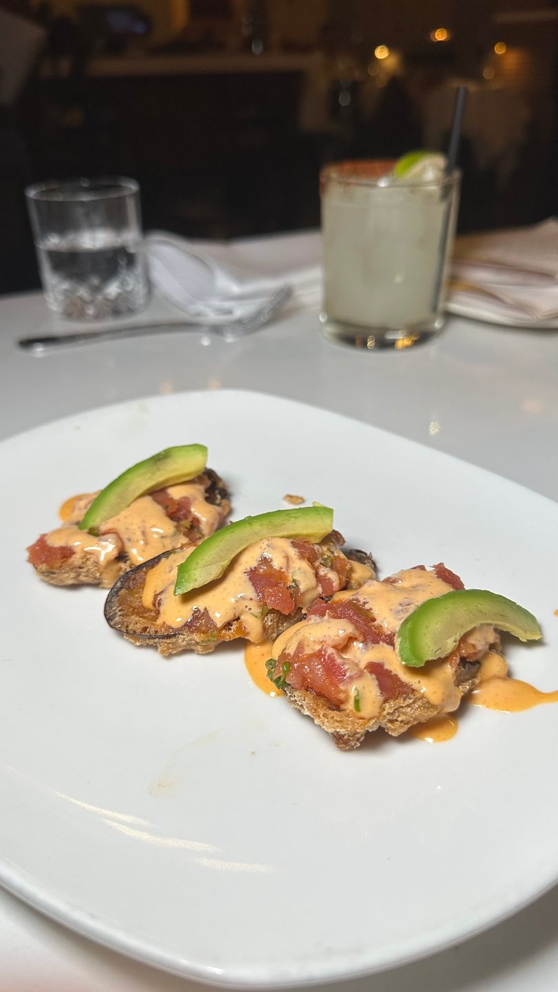Three appetizers with diced tomatoes, creamy sauce, and avocado slices on a white plate, with drinks in the background.